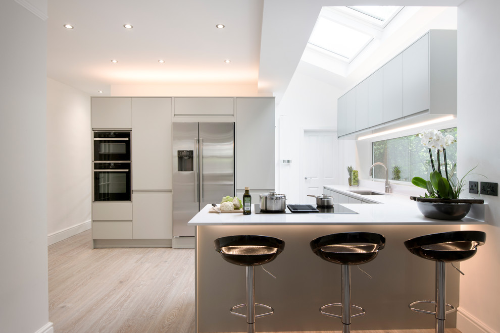 Medium sized contemporary u-shaped open plan kitchen in West Midlands with grey cabinets, light hardwood flooring, a breakfast bar, white worktops, a submerged sink, flat-panel cabinets, window splashback, stainless steel appliances and beige floors.