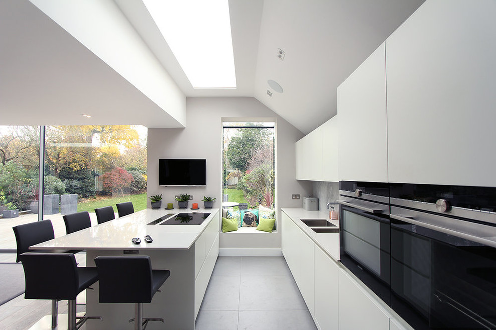 Стильный дизайн: параллельная кухня в современном стиле с двойной мойкой, плоскими фасадами, белыми фасадами, серым фартуком, черной техникой, полуостровом, серым полом, белой столешницей и телевизором - последний тренд