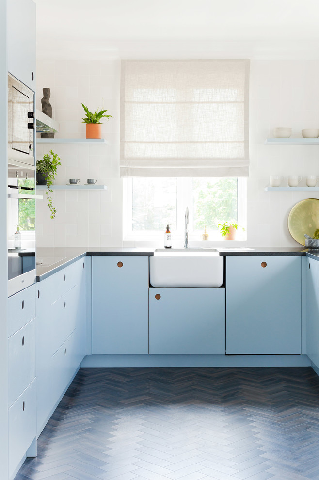 Modern inredning av ett mellanstort u-kök, med en rustik diskho, släta luckor, blå skåp, mörkt trägolv, fönster som stänkskydd och brunt golv