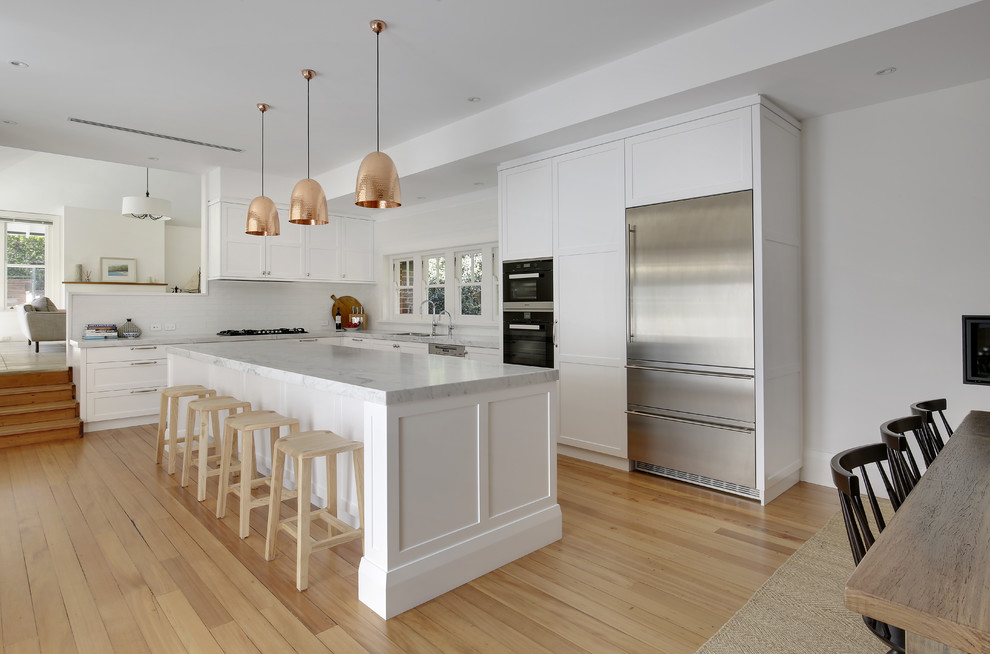 Inspiration for a large timeless l-shaped light wood floor and yellow floor eat-in kitchen remodel in Sydney with a double-bowl sink, shaker cabinets, white cabinets, marble countertops, white backsplash, ceramic backsplash, stainless steel appliances and an island