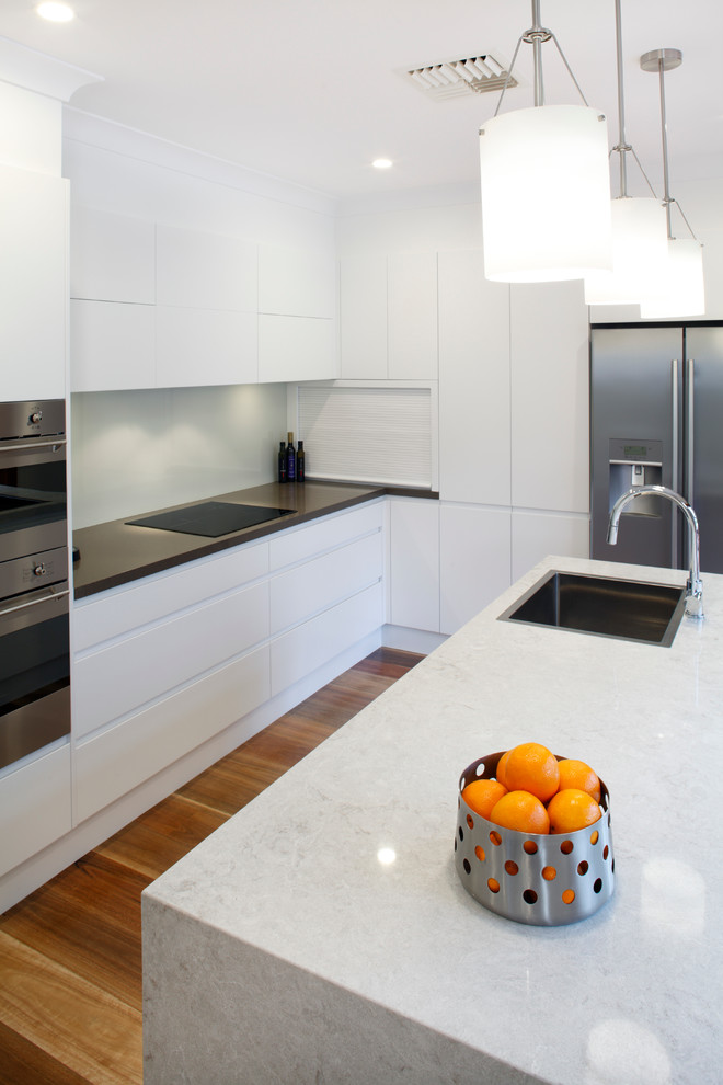 Mittelgroße Moderne Küche in L-Form mit Waschbecken, flächenbündigen Schrankfronten, weißen Schränken, Quarzwerkstein-Arbeitsplatte, Küchenrückwand in Weiß, Glasrückwand, braunem Holzboden und Kücheninsel in Sydney