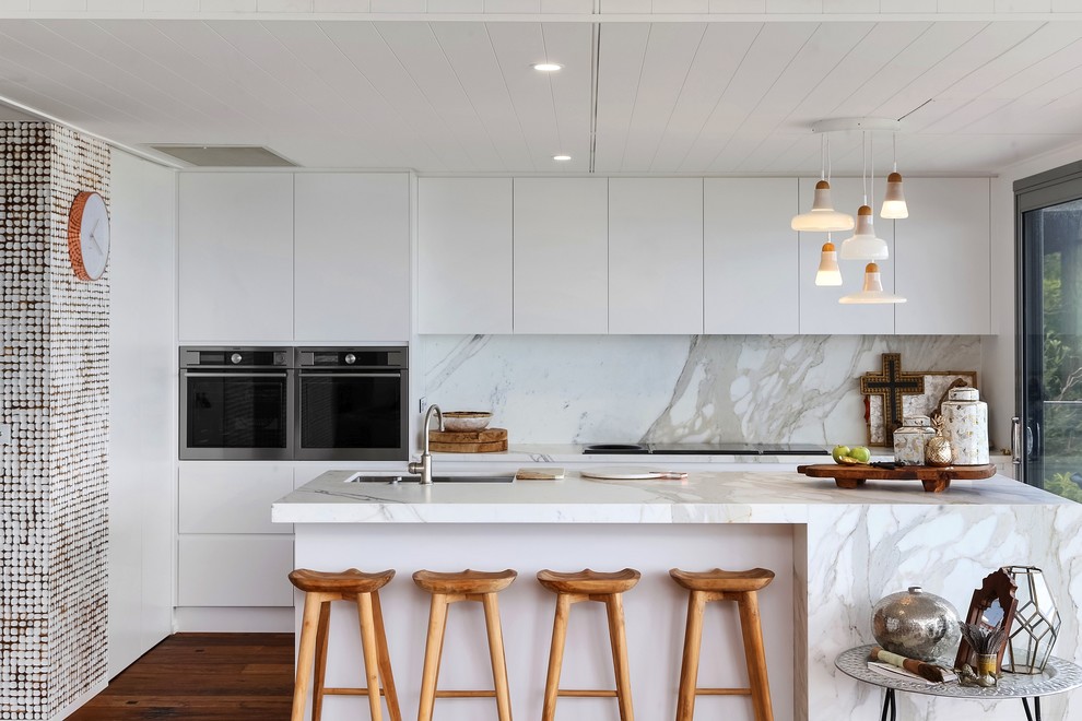 Photo of a world-inspired galley kitchen in Central Coast with a submerged sink, flat-panel cabinets, white cabinets, white splashback, stainless steel appliances, dark hardwood flooring, an island and brown floors.