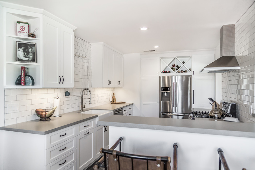 Example of a mid-sized trendy u-shaped laminate floor and black floor enclosed kitchen design in Orange County with a farmhouse sink, shaker cabinets, white cabinets, concrete countertops, white backsplash, subway tile backsplash, stainless steel appliances, a peninsula and gray countertops