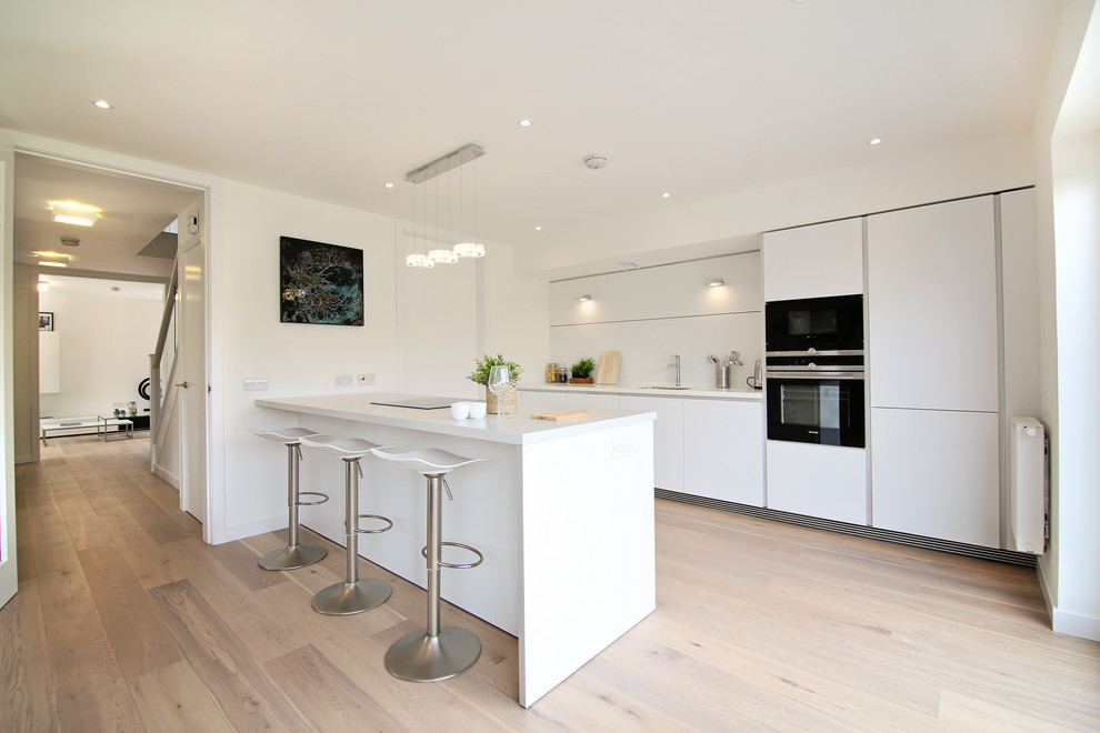 Inspiration for a large contemporary single-wall open plan kitchen in Glasgow with an integrated sink, flat-panel cabinets, white cabinets, composite countertops, white splashback, black appliances, light hardwood flooring, a breakfast bar, white worktops and brown floors.
