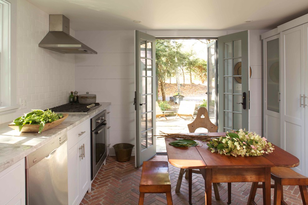 This is an example of a country kitchen in New York with shaker cabinets, white cabinets, marble worktops, white splashback, metro tiled splashback, stainless steel appliances and brick flooring.