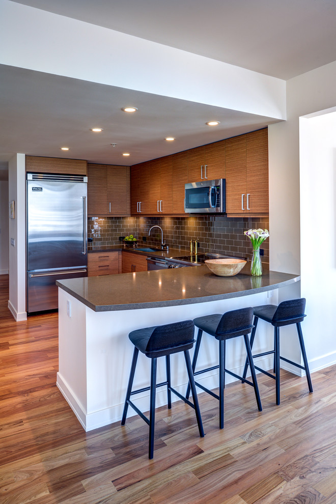 Small modern u-shaped kitchen/diner in San Francisco with a submerged sink, flat-panel cabinets, medium wood cabinets, engineered stone countertops, green splashback, stainless steel appliances, a breakfast bar, glass tiled splashback and medium hardwood flooring.