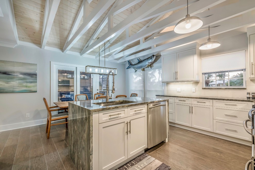 Bild på ett mellanstort maritimt grå grått kök, med en enkel diskho, skåp i shakerstil, vita skåp, bänkskiva i kvartsit, grått stänkskydd, stänkskydd i glaskakel, rostfria vitvaror, mellanmörkt trägolv, en köksö och grått golv