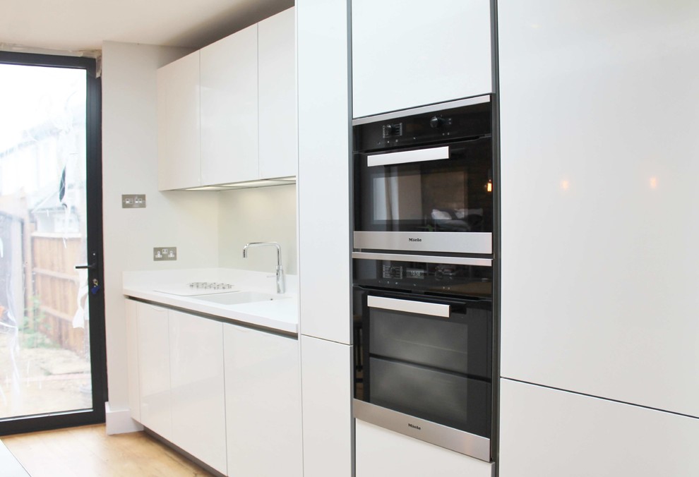 Photo of a small contemporary single-wall kitchen/diner in London with a built-in sink, flat-panel cabinets, white cabinets, concrete worktops, metallic splashback, glass sheet splashback, stainless steel appliances and an island.