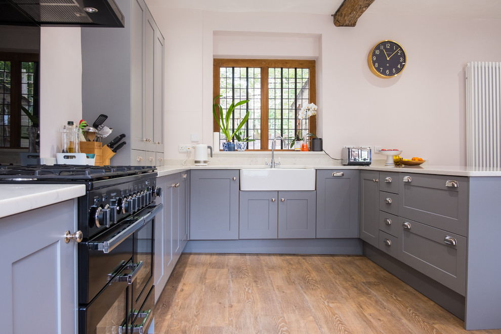 Design ideas for a large classic u-shaped open plan kitchen in Gloucestershire with a belfast sink, shaker cabinets, blue cabinets, quartz worktops, black appliances, no island, white worktops, medium hardwood flooring and brown floors.