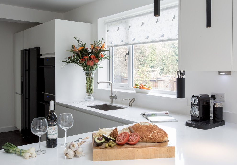 Medium sized contemporary u-shaped open plan kitchen in London with a submerged sink, flat-panel cabinets, white cabinets, composite countertops, black appliances, dark hardwood flooring and a breakfast bar.