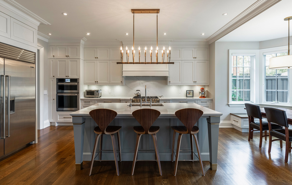 Elegant l-shaped medium tone wood floor and brown floor eat-in kitchen photo in Toronto with an undermount sink, recessed-panel cabinets, gray cabinets, white backsplash, stainless steel appliances, an island and white countertops