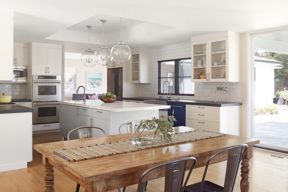 Photo of a coastal open plan kitchen in San Francisco with a belfast sink, glass-front cabinets, white cabinets, white splashback, metro tiled splashback, stainless steel appliances, light hardwood flooring, an island, beige floors and grey worktops.