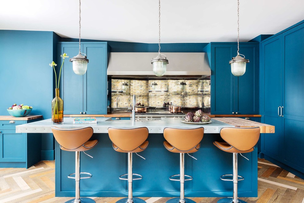 Photo of a classic l-shaped kitchen/diner in London with a belfast sink, shaker cabinets, blue cabinets, metallic splashback, mirror splashback, integrated appliances, medium hardwood flooring, an island, beige floors and grey worktops.