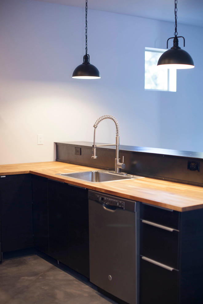 Foto på ett litet industriellt kök, med en enkel diskho, släta luckor, svarta skåp, träbänkskiva, svart stänkskydd och betonggolv