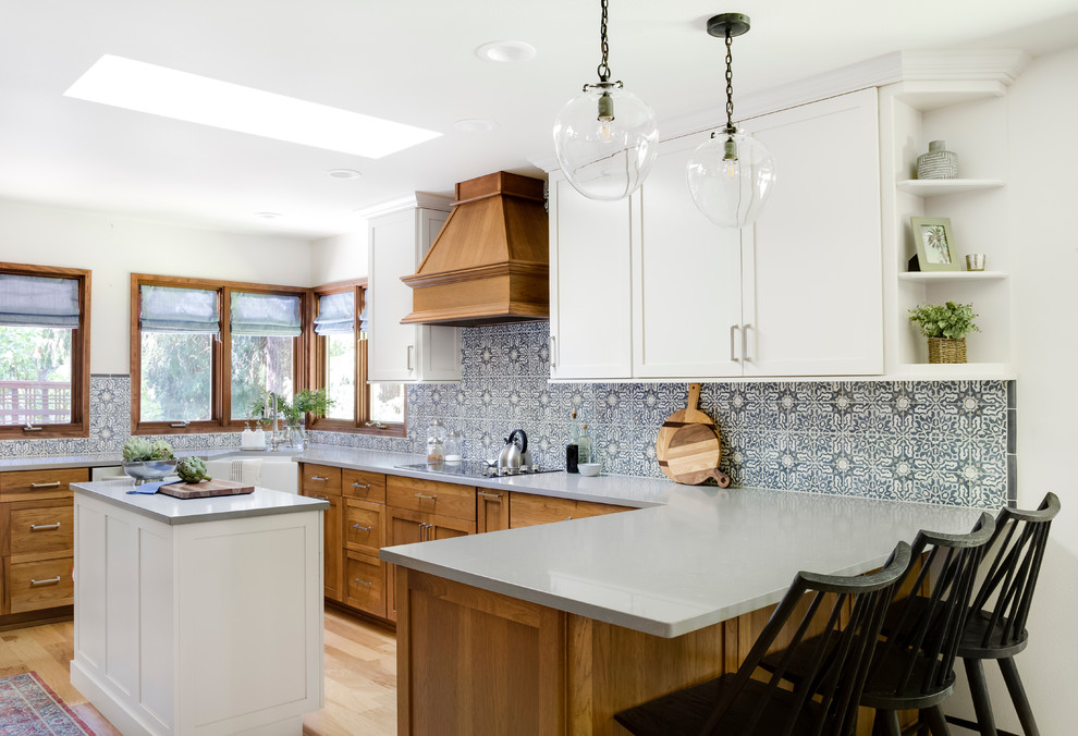 Photo of a medium sized farmhouse u-shaped kitchen/diner in Seattle with a belfast sink, shaker cabinets, white cabinets, engineered stone countertops, multi-coloured splashback, ceramic splashback, stainless steel appliances, light hardwood flooring, an island, brown floors and grey worktops.