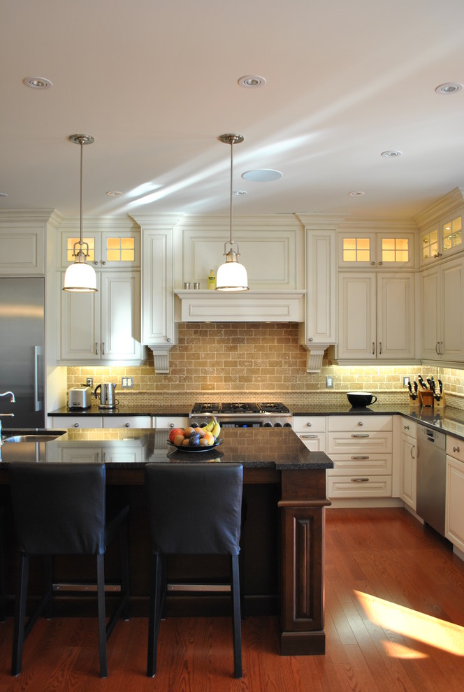 Cette photo montre une cuisine bicolore chic en L avec un placard avec porte à panneau surélevé, un électroménager en acier inoxydable, des portes de placard blanches et une crédence en travertin.