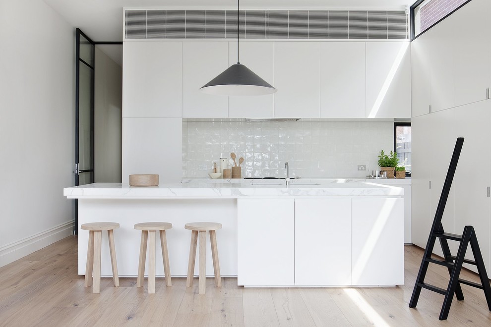 Example of a minimalist l-shaped light wood floor and beige floor kitchen design in Los Angeles with an undermount sink, flat-panel cabinets, white cabinets, white backsplash, paneled appliances, an island and white countertops