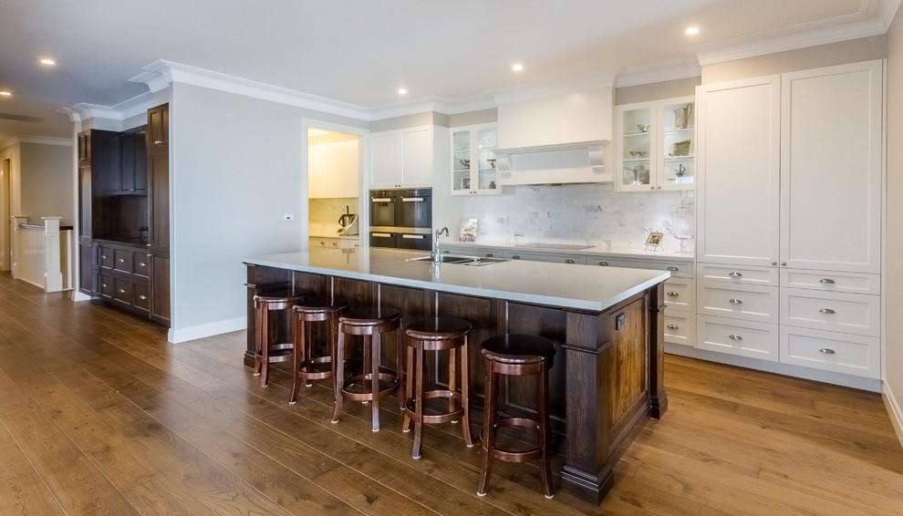 Example of a large classic galley light wood floor open concept kitchen design in Canberra - Queanbeyan with an undermount sink, recessed-panel cabinets, dark wood cabinets, solid surface countertops, beige backsplash, stone tile backsplash, stainless steel appliances and an island