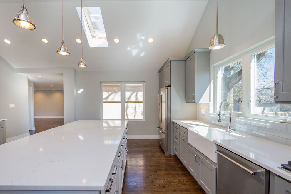 Large contemporary l-shaped kitchen in Chicago with a built-in sink, shaker cabinets, grey cabinets, engineered stone countertops, white splashback, metro tiled splashback, stainless steel appliances, dark hardwood flooring, brown floors and white worktops.