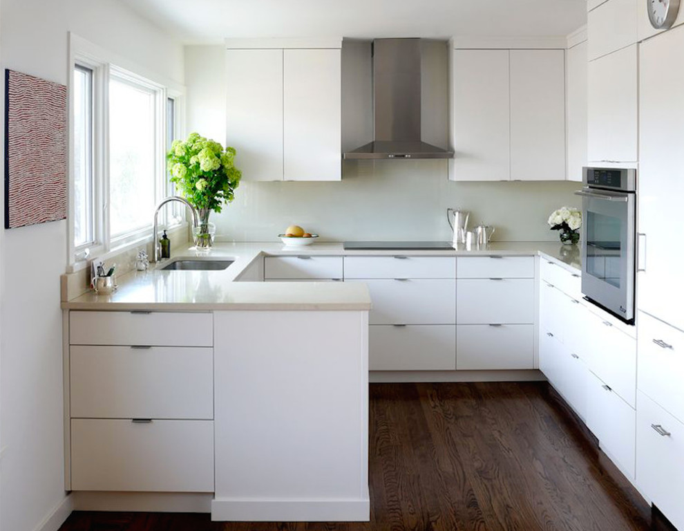 Medium sized traditional u-shaped kitchen/diner in New York with a submerged sink, flat-panel cabinets, white cabinets, composite countertops, white splashback, glass sheet splashback, stainless steel appliances, dark hardwood flooring and brown floors.