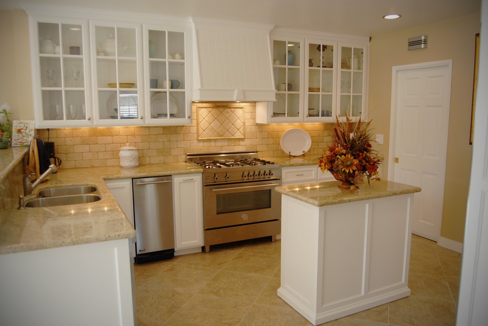Mid-sized elegant l-shaped ceramic tile and beige floor enclosed kitchen photo in Orange County with a double-bowl sink, glass-front cabinets, white cabinets, limestone countertops, beige backsplash, stone tile backsplash, stainless steel appliances and a peninsula