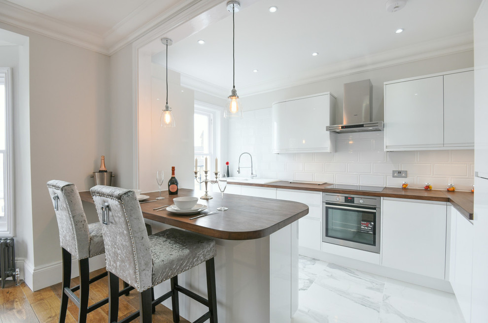 Medium sized contemporary l-shaped open plan kitchen in London with a double-bowl sink, flat-panel cabinets, white cabinets, wood worktops, white splashback, porcelain splashback, integrated appliances, porcelain flooring and a breakfast bar.