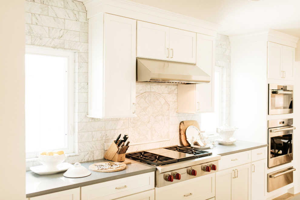 Klassisk inredning av ett mellanstort grå grått kök, med en undermonterad diskho, luckor med infälld panel, vita skåp, bänkskiva i kvarts, vitt stänkskydd, stänkskydd i porslinskakel, rostfria vitvaror, ljust trägolv, en köksö och beiget golv
