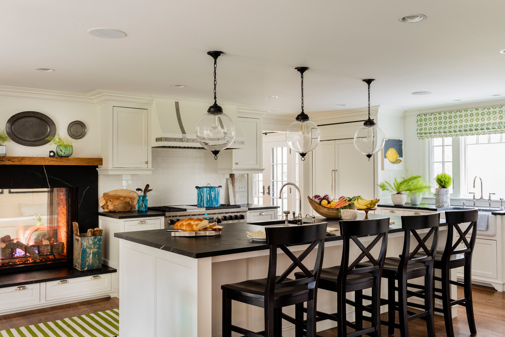 Large transitional l-shaped medium tone wood floor and brown floor eat-in kitchen photo in Boston with a farmhouse sink, flat-panel cabinets, white cabinets, soapstone countertops, white backsplash, subway tile backsplash, paneled appliances and an island
