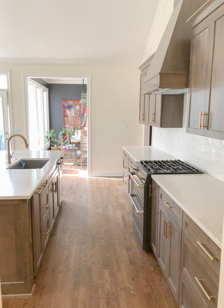 This is an example of a medium sized traditional galley open plan kitchen in Kansas City with a single-bowl sink, shaker cabinets, brown cabinets, engineered stone countertops, white splashback, ceramic splashback, black appliances, medium hardwood flooring, an island, brown floors and white worktops.