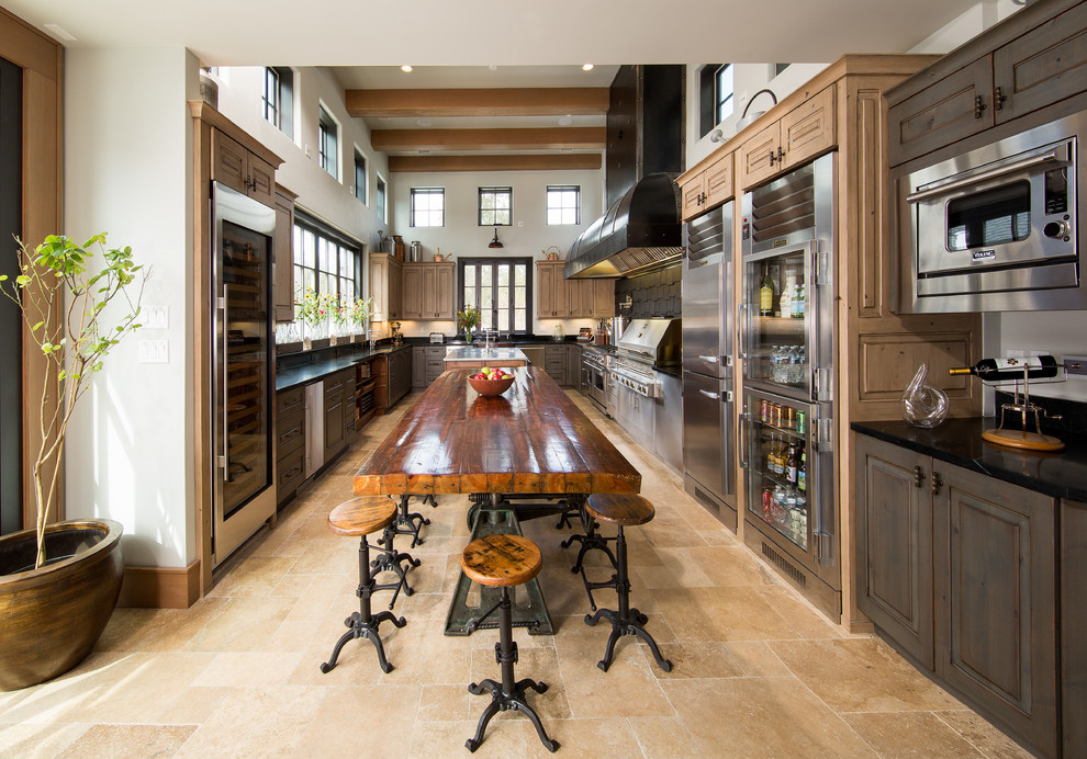 Immagine di una cucina country con ante con bugna sagomata, ante in legno bruno, elettrodomestici in acciaio inossidabile, pavimento beige e top nero