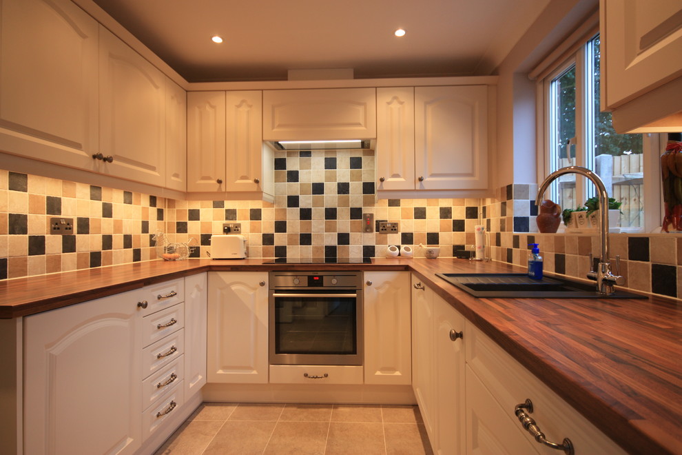This is an example of a medium sized country u-shaped enclosed kitchen in Kent with a built-in sink, recessed-panel cabinets, beige cabinets, wood worktops, multi-coloured splashback, ceramic splashback, stainless steel appliances, ceramic flooring and no island.