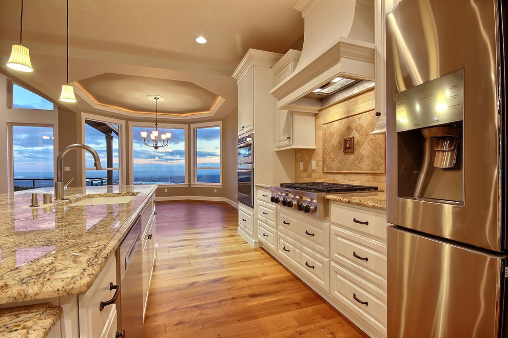 Example of a huge transitional l-shaped medium tone wood floor and brown floor open concept kitchen design in Portland with raised-panel cabinets, white cabinets, granite countertops, brown backsplash, travertine backsplash, stainless steel appliances, an island and an undermount sink