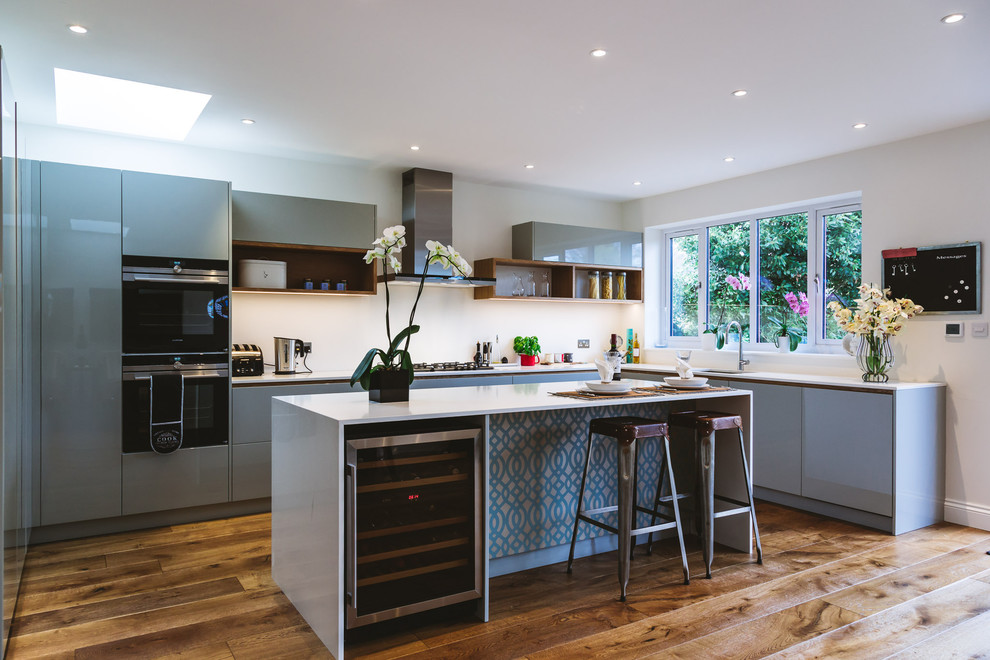 This is an example of a medium sized contemporary u-shaped kitchen in Surrey with flat-panel cabinets, grey cabinets, quartz worktops, stainless steel appliances, light hardwood flooring, an island, beige floors and white worktops.