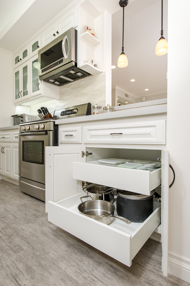 This is an example of a small classic galley enclosed kitchen in Los Angeles with a submerged sink, glass-front cabinets, white cabinets, engineered stone countertops, beige splashback, ceramic splashback, stainless steel appliances and porcelain flooring.