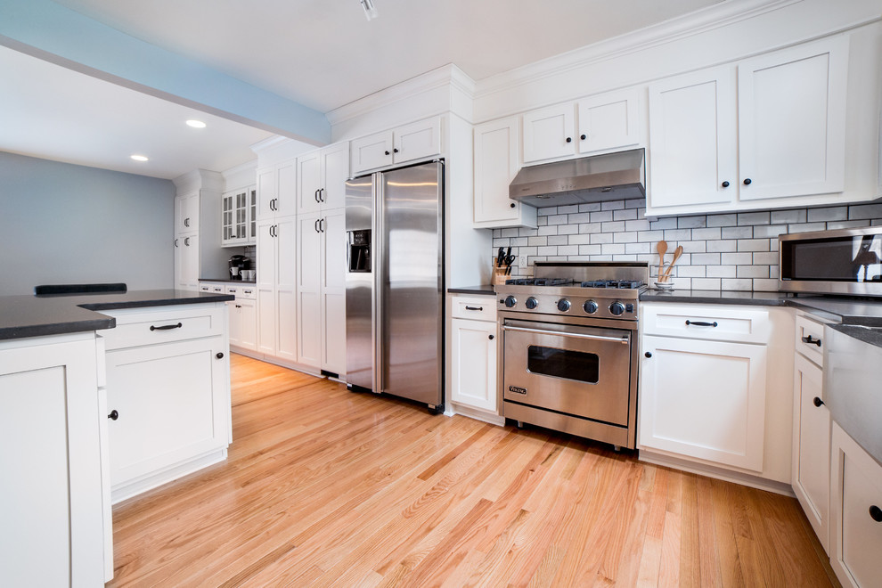 Small transitional l-shaped light wood floor open concept kitchen photo in New York with a farmhouse sink, shaker cabinets, white cabinets, quartz countertops, white backsplash, subway tile backsplash, stainless steel appliances and a peninsula