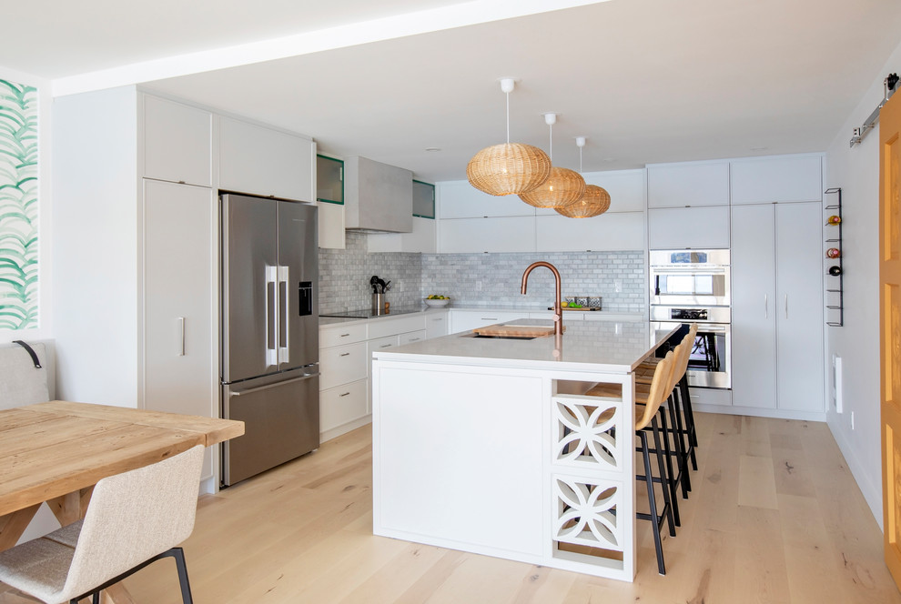 World-inspired l-shaped kitchen/diner in Other with a submerged sink, flat-panel cabinets, white cabinets, grey splashback, metro tiled splashback, stainless steel appliances, light hardwood flooring, an island and white worktops.