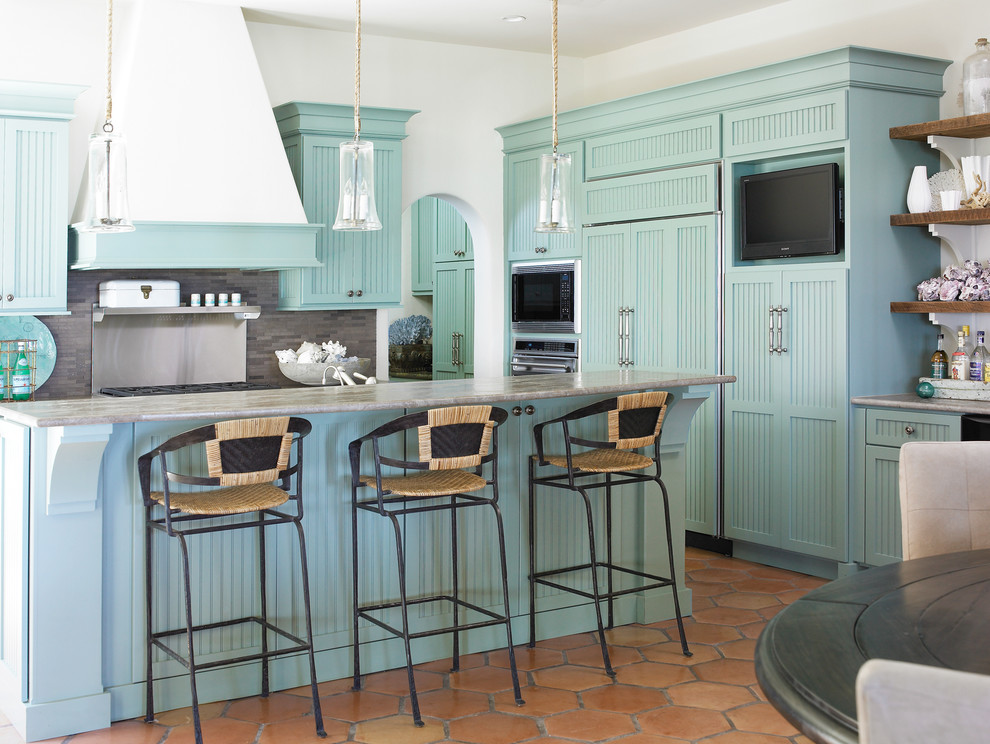 На фото: угловая кухня в морском стиле с обеденным столом, синими фасадами, серым фартуком и техникой под мебельный фасад