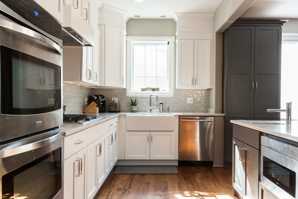 This is an example of a large classic l-shaped kitchen/diner in Chicago with a belfast sink, shaker cabinets, white cabinets, engineered stone countertops, grey splashback, ceramic splashback, stainless steel appliances, medium hardwood flooring, an island, brown floors and grey worktops.