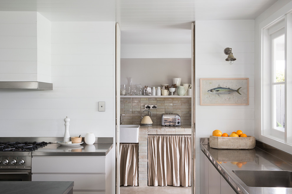 Imagen de cocina costera con fregadero integrado, armarios con paneles lisos, puertas de armario blancas, encimera de acero inoxidable y electrodomésticos de acero inoxidable