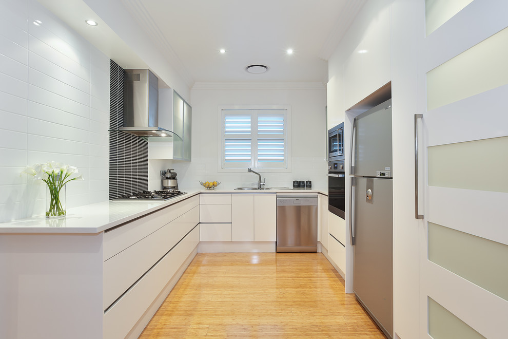 Example of a mid-sized trendy u-shaped bamboo floor and beige floor enclosed kitchen design in Sydney with a drop-in sink, flat-panel cabinets, white cabinets, white backsplash, stainless steel appliances, quartz countertops, ceramic backsplash and white countertops
