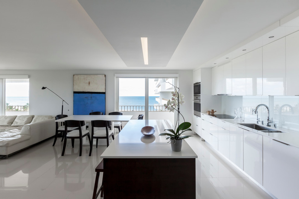 Свежая идея для дизайна: кухня-гостиная в современном стиле с врезной мойкой, плоскими фасадами, белыми фасадами, белым фартуком, фартуком из стекла, техникой из нержавеющей стали и островом - отличное фото интерьера
