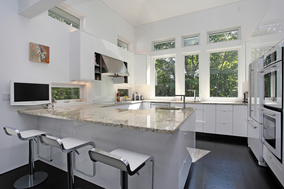 This is an example of a contemporary u-shaped open plan kitchen in New York with flat-panel cabinets, white cabinets, glass sheet splashback, white appliances, white splashback, granite worktops and dark hardwood flooring.