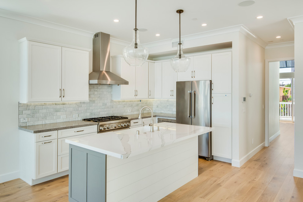 Medium sized traditional l-shaped kitchen/diner in Los Angeles with a belfast sink, shaker cabinets, grey cabinets, engineered stone countertops, grey splashback, stone tiled splashback, stainless steel appliances, light hardwood flooring and an island.