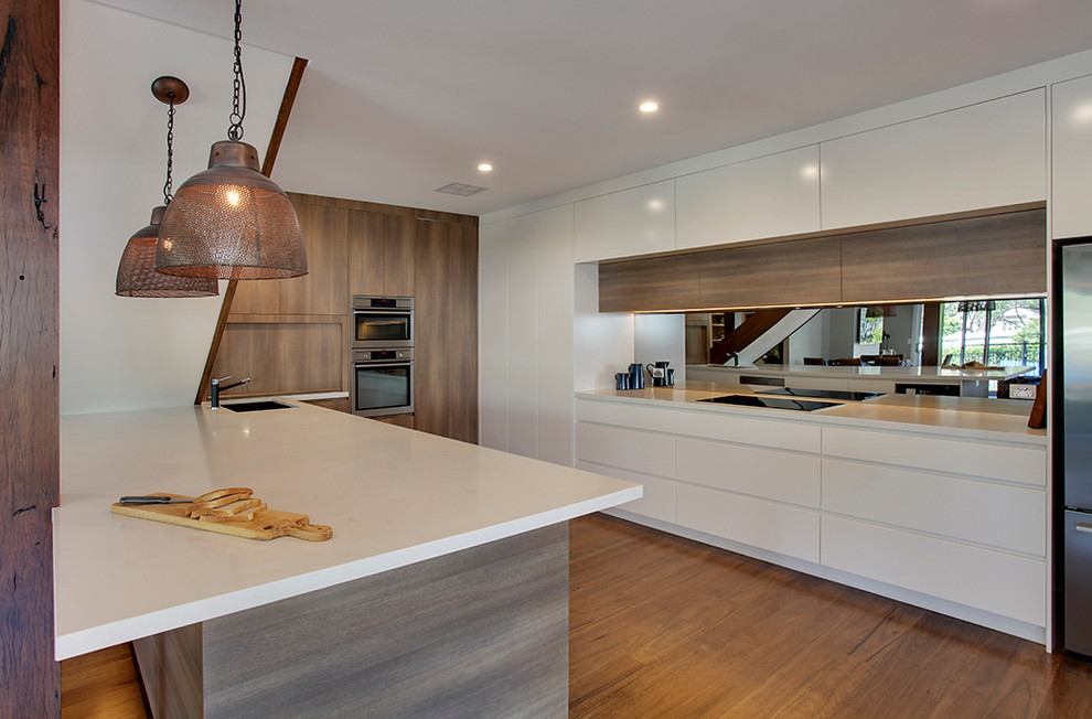 Large contemporary galley open plan kitchen in Sydney with a submerged sink, flat-panel cabinets, white cabinets, engineered stone countertops, metallic splashback, mirror splashback, stainless steel appliances, light hardwood flooring and an island.