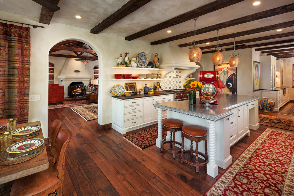 Cette image montre une grande cuisine américaine méditerranéenne avec des portes de placard blanches, une crédence multicolore, un électroménager en acier inoxydable, îlot, un placard sans porte, un sol en bois brun et un sol orange.