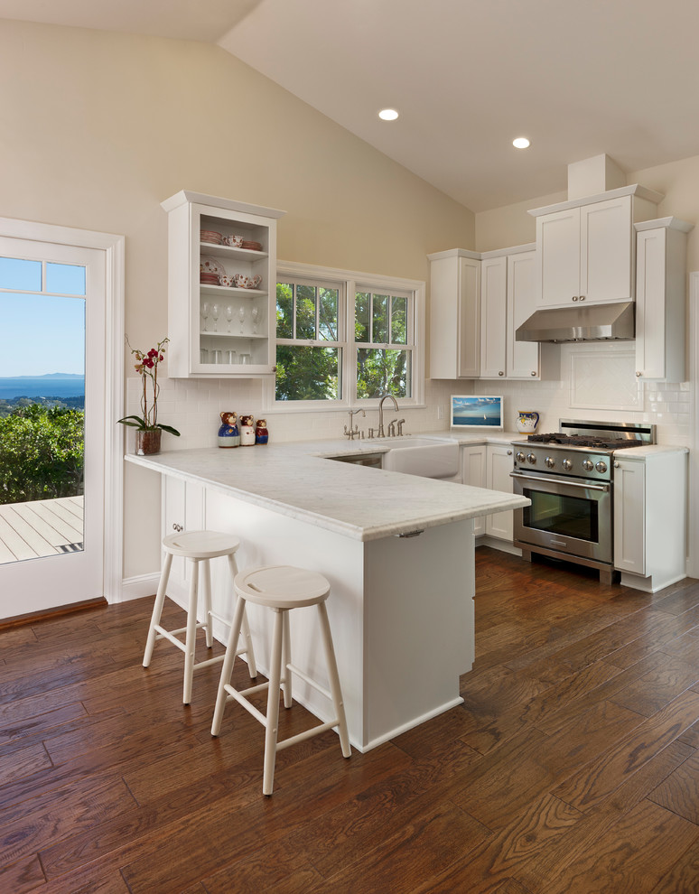 Inspiration for a small l-shaped medium tone wood floor open concept kitchen remodel in Santa Barbara with a farmhouse sink, recessed-panel cabinets, white cabinets, marble countertops, white backsplash, subway tile backsplash, stainless steel appliances and a peninsula