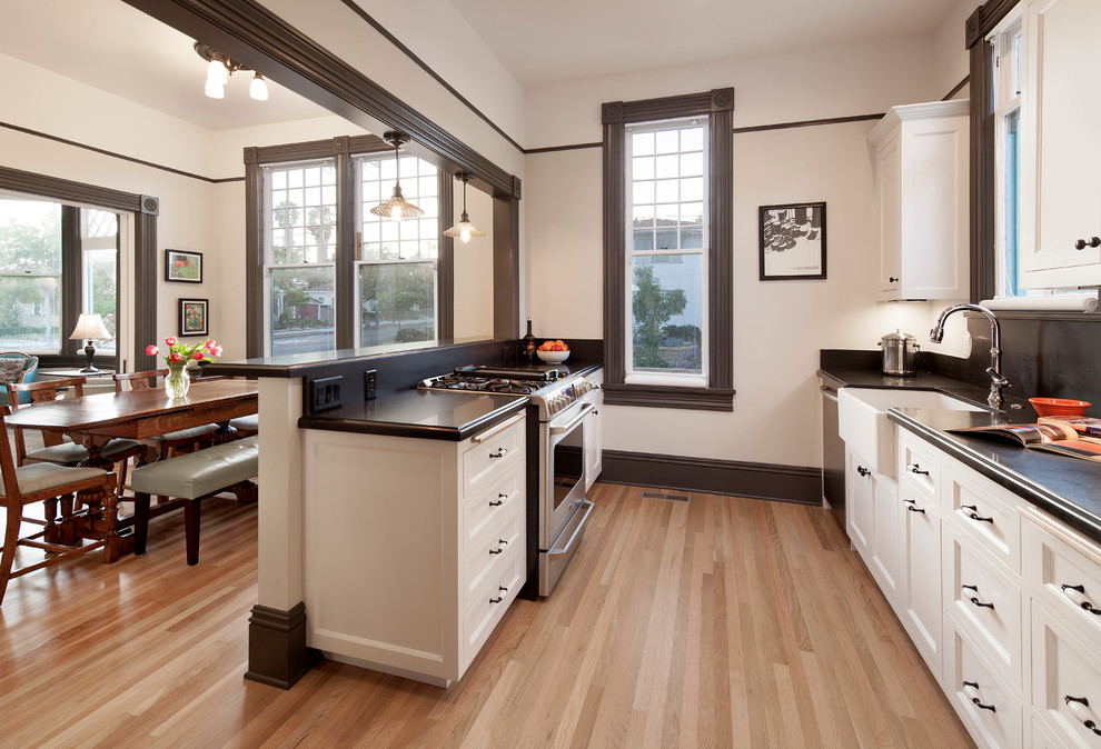 Inspiration for a small victorian galley kitchen/diner in Santa Barbara with a belfast sink, recessed-panel cabinets, white cabinets, engineered stone countertops, black splashback, stainless steel appliances, light hardwood flooring, no island and beige floors.