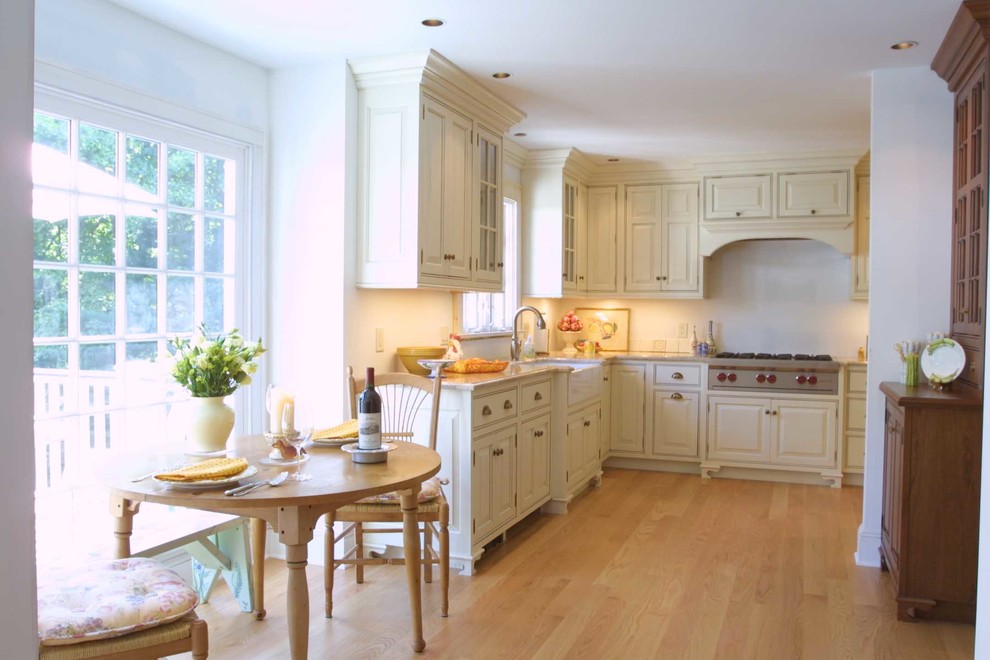 This is an example of a traditional l-shaped kitchen/diner in New York with a belfast sink, beaded cabinets, white cabinets, granite worktops, beige splashback, stainless steel appliances and light hardwood flooring.