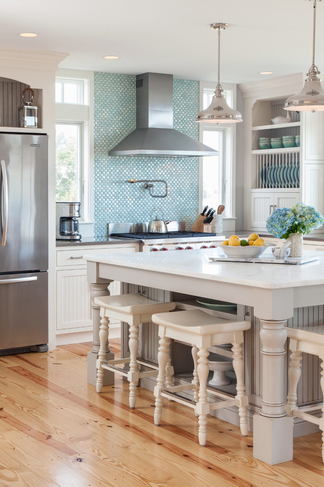 This is an example of a large beach style l-shaped kitchen/diner in Boston with recessed-panel cabinets, white cabinets, blue splashback, mosaic tiled splashback, stainless steel appliances, light hardwood flooring, an island and composite countertops.