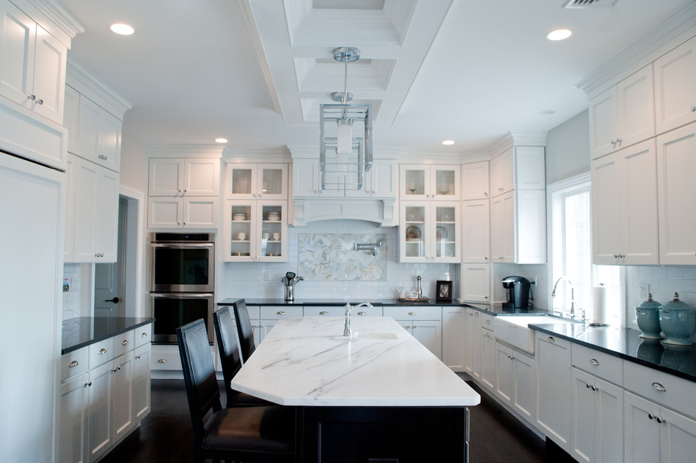 Photo of a large contemporary u-shaped kitchen/diner in New York with a belfast sink, recessed-panel cabinets, white cabinets, granite worktops, white splashback, metro tiled splashback, stainless steel appliances, dark hardwood flooring, an island, brown floors, black worktops and a coffered ceiling.